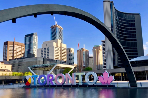 The Toronto Sign at City Hall stands at the edge of a pool on a sunny day. City Hall and the Toronto skyline are visible in the background.