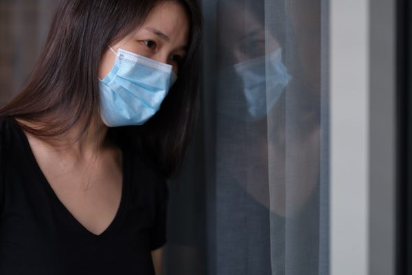 A woman leans against a window while wearing a medical mask. Her expression is tired and sad.