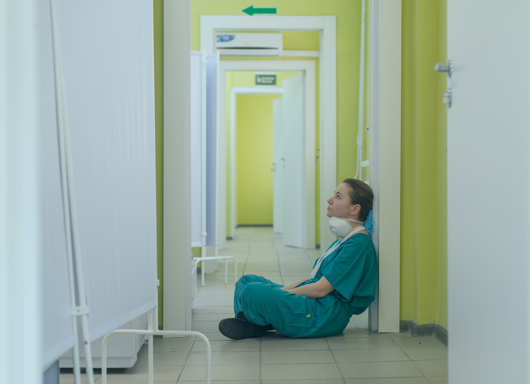 A health care worker in scrubs sits on the floor in a hospital corridor, looking exhausted